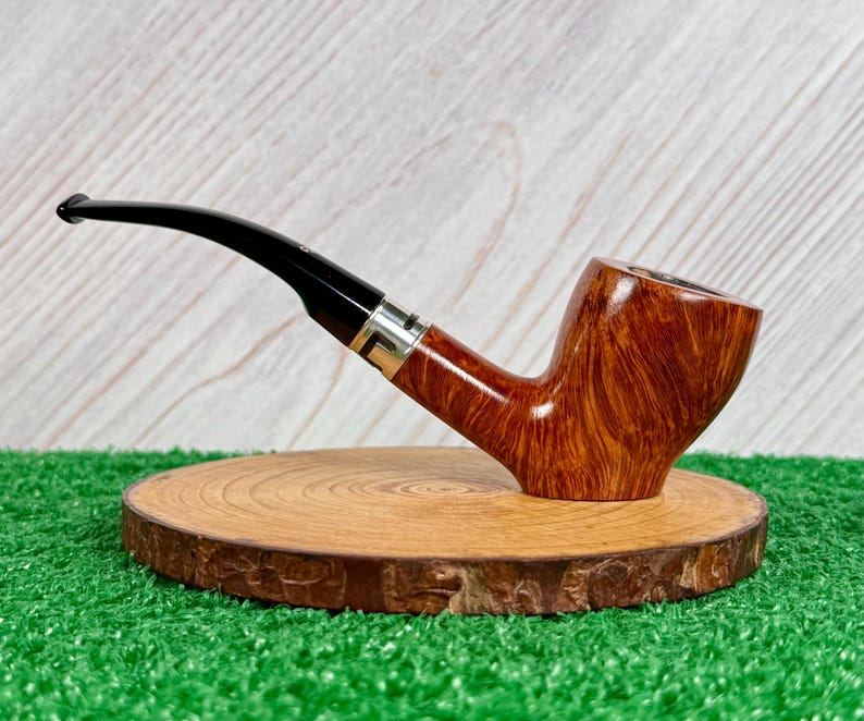 May include: A tobacco pipe with a polished brown wood grain bowl and a curved black stem. The stem features a silver-colored metal band where it connects to the bowl. The pipe is positioned horizontally, showcasing the smooth finish and natural wood texture.