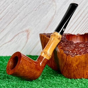 May include: A handcrafted tobacco pipe with a curved, brown wooden bowl and a bamboo stem. The stem has a black mouthpiece and a decorative bamboo section. The pipe rests on a green surface next to a wooden bowl.