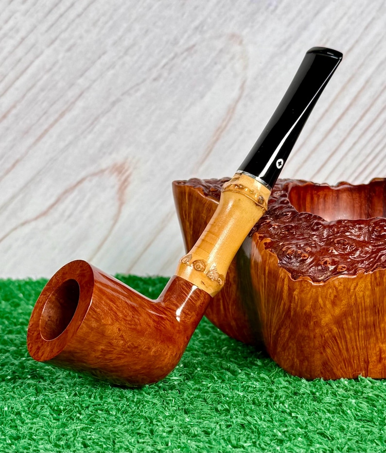 May include: A handcrafted tobacco pipe with a curved, brown wooden bowl and a bamboo stem. The stem has a black mouthpiece and a decorative bamboo section. The pipe rests on a green surface next to a wooden bowl.