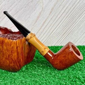 May include: A tobacco pipe with a dark brown bowl and a light brown bamboo-style stem. The mouthpiece is black. The pipe rests on a green surface. The bowl is next to a wooden container.
