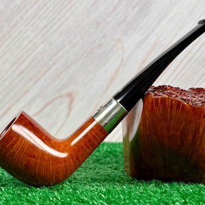 Pode incluir: Um cachimbo de tabaco com fornilho de madeira polida castanho-claro e uma piteira preta longa e fina. Uma banda de metal prateado liga o fornilho à piteira. O cachimbo está posicionado ao lado de um grande pedaço de madeira bruta texturizada.