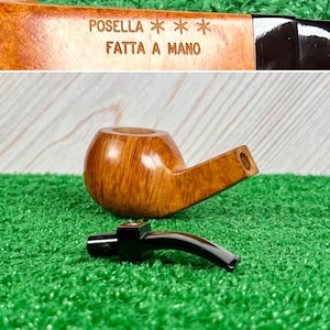 May include: A light brown wooden tobacco pipe with a smooth finish and a detachable black stem. The bowl is engraved with the text POSELLA *** FATTA A MANO. The pipe is shown in two parts, with the bowl and the stem separated on a green surface.