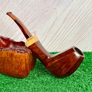 Pode incluir: Um cachimbo de tabaco de madeira castanha polida com uma haste curva e lisa e uma faixa de madeira clara na jun&ccedil;&atilde;o. O cachimbo apresenta um padr&atilde;o de gr&atilde;o profundo e rico e um acabamento escuro e brilhante, apoiado num bloco claro.