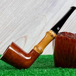 May include: A handcrafted tobacco pipe with a dark brown bowl and a light brown bamboo stem. The stem has a black mouthpiece with a white accent. The pipe rests on a textured wooden surface and green turf.