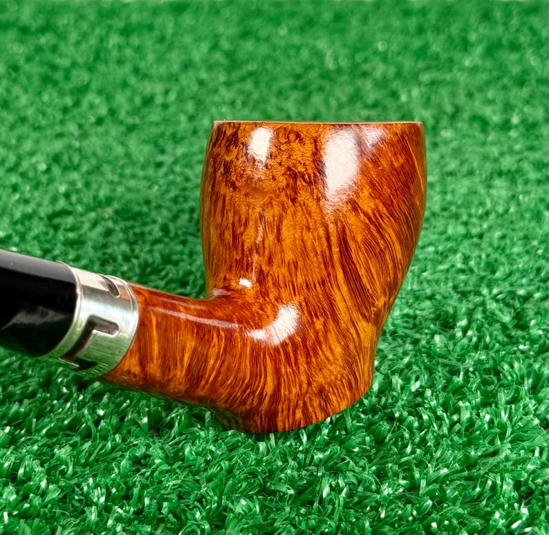 May include: A tobacco pipe featuring a polished, light brown wooden bowl with a prominent grain pattern. The stem is black and connects to the bowl via a silver-colored metal band. The pipe has a smooth, glossy finish and a curved shape.