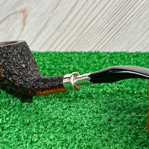May include: A tobacco pipe with a dark, textured bowl and a black, curved stem. The pipe features a silver band and rests on a green surface. The bowl has a rough, bark-like texture.