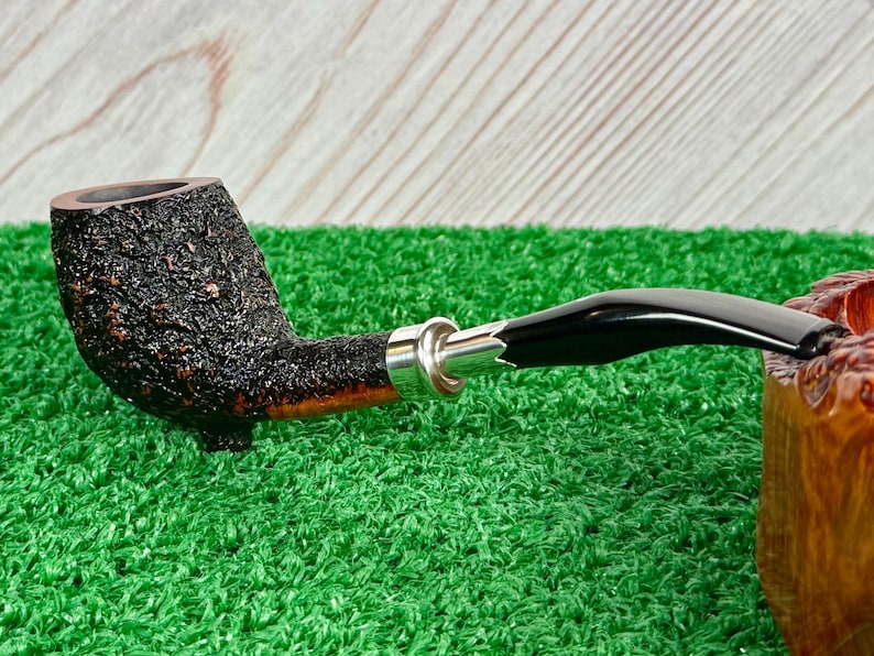 May include: A tobacco pipe with a dark, textured bowl and a black, curved stem. The pipe features a silver band and rests on a green surface. The bowl has a rough, bark-like texture.