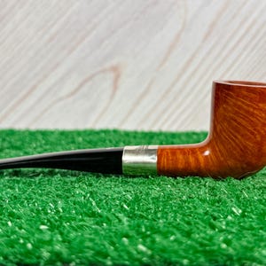 Puede incluir: Una pipa de tabaco con un hornillo de madera marr&oacute;n pulida con veta visible, una boquilla negra lisa y una banda de metal plateado que conecta ambas secciones. La pipa est&aacute; colocada horizontalmente sobre una superficie texturizada.