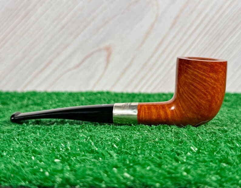 Puede incluir: Una pipa de tabaco con un hornillo de madera marr&oacute;n pulida con veta visible, una boquilla negra lisa y una banda de metal plateado que conecta ambas secciones. La pipa est&aacute; colocada horizontalmente sobre una superficie texturizada.
