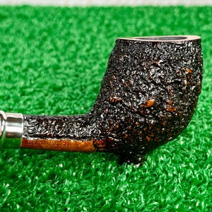 May include: Close-up of a tobacco pipe with a dark, textured bowl and a silver band. The stem is a lighter brown color. The pipe is set against a green background.