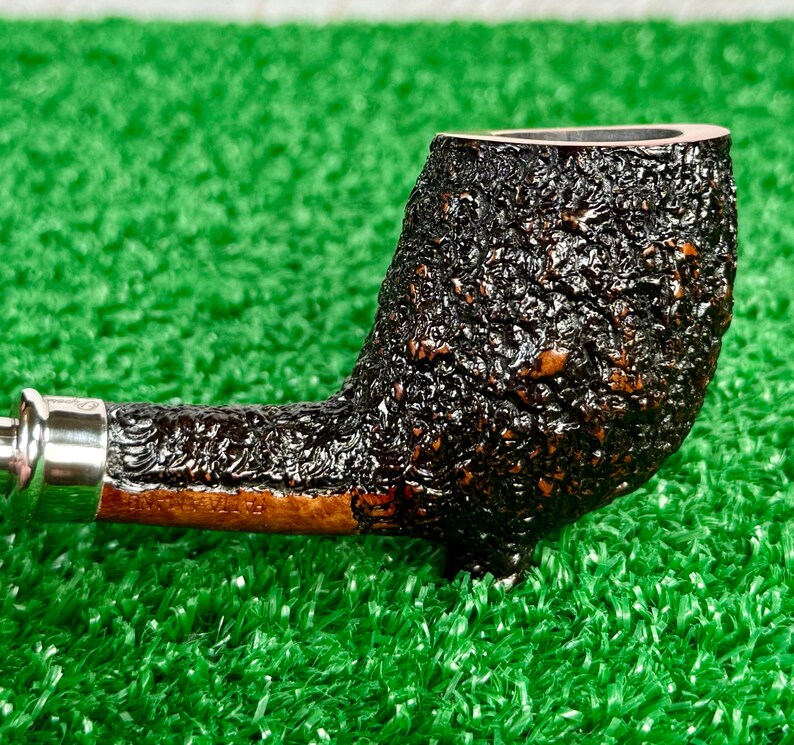 May include: Close-up of a tobacco pipe with a dark, textured bowl and a silver band. The stem is a lighter brown color. The pipe is set against a green background.