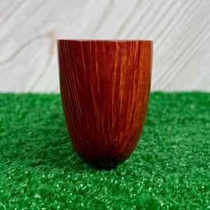 May include: A smooth, cylindrical wooden vessel with a rich, reddish-brown finish and prominent vertical grain patterns. The object features a wide, open top and a rounded, tapered base, showcasing the natural texture and warm tones of the wood.
