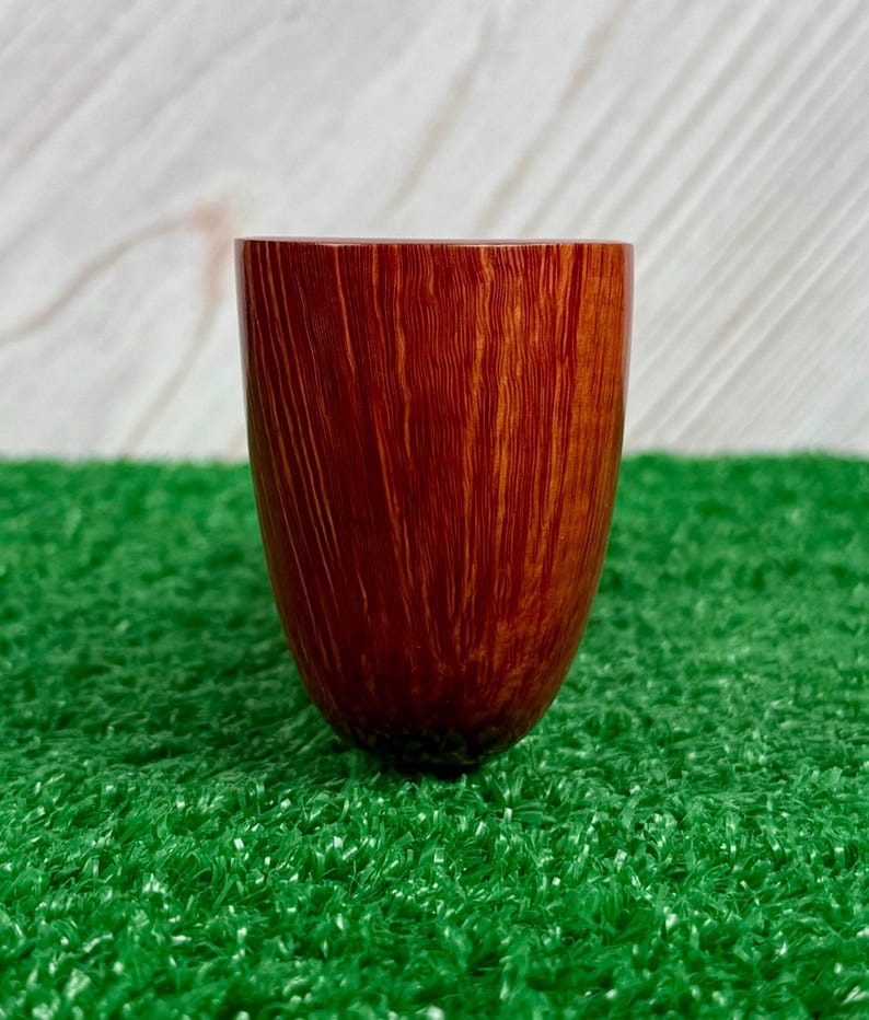 May include: A smooth, cylindrical wooden vessel with a rich, reddish-brown finish and prominent vertical grain patterns. The object features a wide, open top and a rounded, tapered base, showcasing the natural texture and warm tones of the wood.