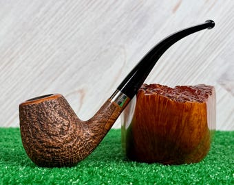 May include: A tobacco pipe with a textured brown briar bowl and a smooth black stem. The pipe features a silver-colored metal band connecting the bowl to the stem. It is positioned next to a polished, light brown wooden block with a natural grain.