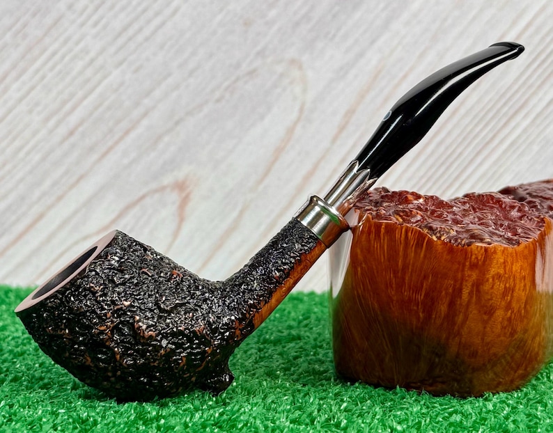 May include: A tobacco pipe with a dark, textured bowl and a black, curved stem. The pipe has a silver band and rests on a wooden block. The pipe is set against a green surface.