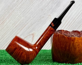May include: A tobacco pipe with a polished, reddish-brown wooden bowl featuring a visible grain pattern. The pipe has a long, slender black stem with a dark, multi-colored accent ring near the bowl connection, resting against a textured wood block.