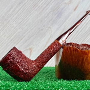 May include: A tobacco pipe with a dark reddish-brown, deeply textured bowl and shank. The stem is smooth, glossy, and dark brown, featuring a small gold-colored letter E logo near the junction with the shank. The pipe is positioned upright.