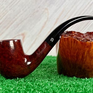 Pode incluir: Um cachimbo de tabaco curvo, castanho, com boquilha preta, repousa sobre uma superfície verde. O cachimbo tem um acabamento brilhante e a palavra "Davidoff" está gravada na lateral. Um bloco de madeira está ao lado do cachimbo.