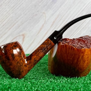 Pode incluir: Um cachimbo de tabaco com um bocal preto curvo e uma piteira castanha polida. A piteira apresenta um padrão de grão rodopiante. O cachimbo repousa sobre um bloco de madeira com superfície texturizada, sobre um fundo verde.