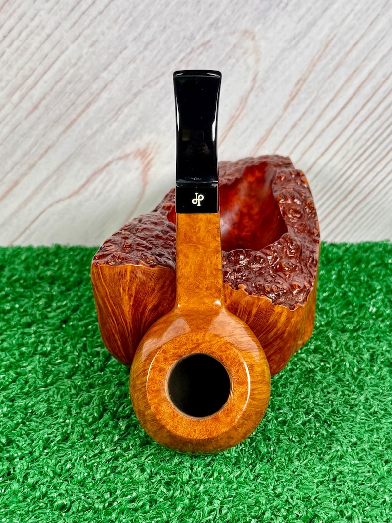 May include: A tobacco pipe with a smooth, light brown wooden bowl and a black stem featuring a white logo with the letters dp. The pipe is positioned in front of a larger, rough-textured, dark brown wooden block with a natural, organic grain pattern.
