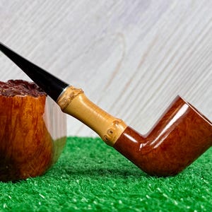 May include: A handcrafted tobacco pipe with a polished, dark brown bowl and a light brown bamboo stem. The stem has a black mouthpiece and is decorated with carved details. The pipe rests on a green surface.
