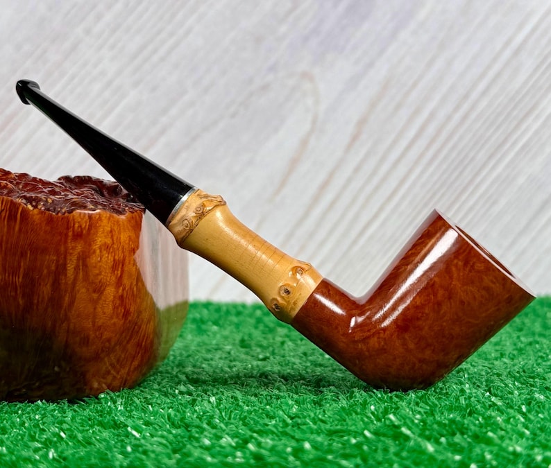 May include: A handcrafted tobacco pipe with a polished, dark brown bowl and a light brown bamboo stem. The stem has a black mouthpiece and is decorated with carved details. The pipe rests on a green surface.
