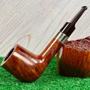 Pode incluir: Um cachimbo de tabaco castanho curvo com um bocal castanho escuro e uma faixa prateada. O cachimbo repousa sobre uma superfície verde junto a uma tigela de madeira. As palavras "NORTHERN BRIAR PREMIER" estão gravadas no cachimbo.