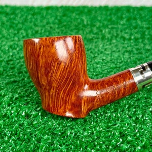May include: A polished brown tobacco pipe with a prominent wood grain pattern and a smooth, glossy finish. The pipe features a silver-colored metal band at the base of the stem, which is engraved with cursive text that is partially visible.