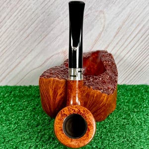 May include: A tobacco pipe with a polished brown burl wood bowl featuring a textured, rugged exterior and a smooth, circular rim. The stem is black with a silver-colored metal band near the junction, and a small white logo is visible on the stem.
