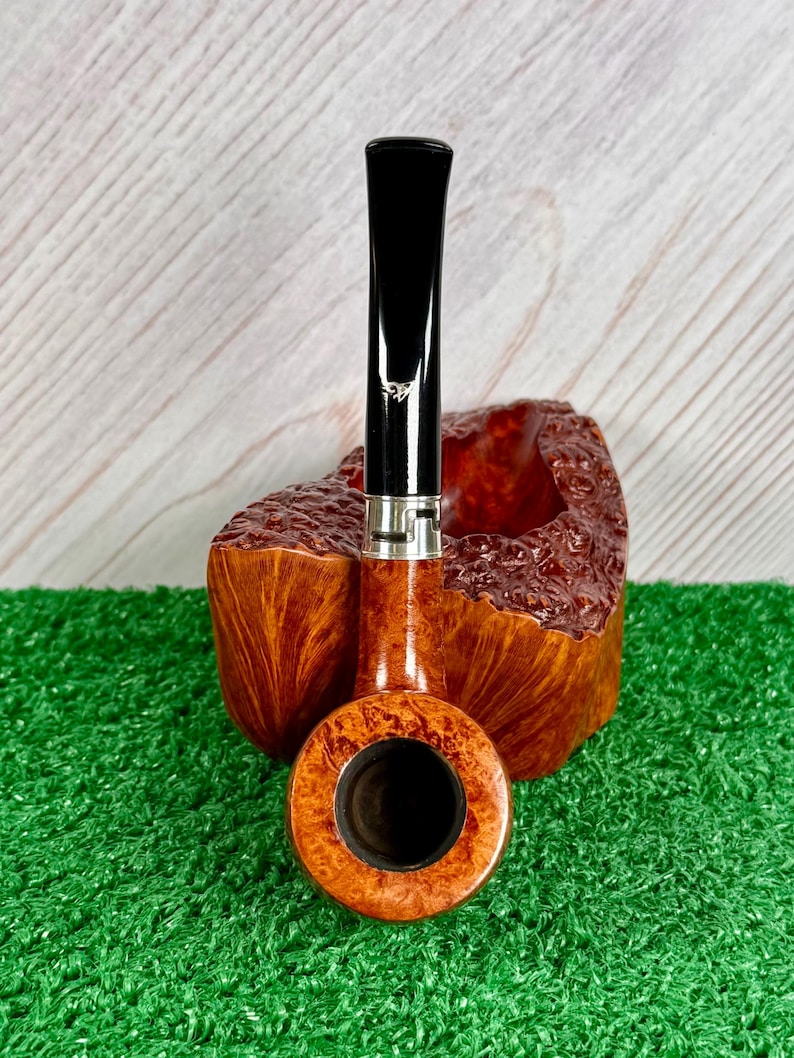 May include: A tobacco pipe with a polished brown burl wood bowl featuring a textured, rugged exterior and a smooth, circular rim. The stem is black with a silver-colored metal band near the junction, and a small white logo is visible on the stem.