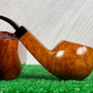 May include: A tobacco pipe featuring a polished light brown wooden bowl with a visible grain pattern and a smooth, curved black stem. The pipe is positioned horizontally, showcasing the contrast between the natural wood finish and the dark stem.
