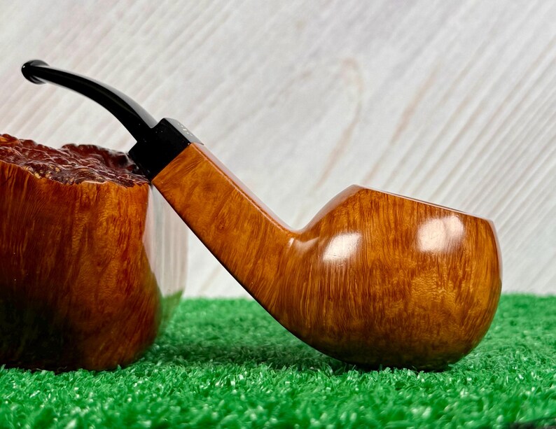 May include: A tobacco pipe featuring a polished light brown wooden bowl with a visible grain pattern and a smooth, curved black stem. The pipe is positioned horizontally, showcasing the contrast between the natural wood finish and the dark stem.