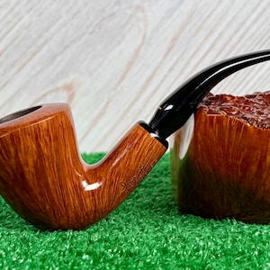 Pode incluir: Um cachimbo de tabaco castanho e curvo com boquilha preta, repousando sobre uma superfície verde. O cachimbo tem um acabamento polido, mostrando o grão da madeira. Um suporte de cachimbo de madeira é visível no fundo.