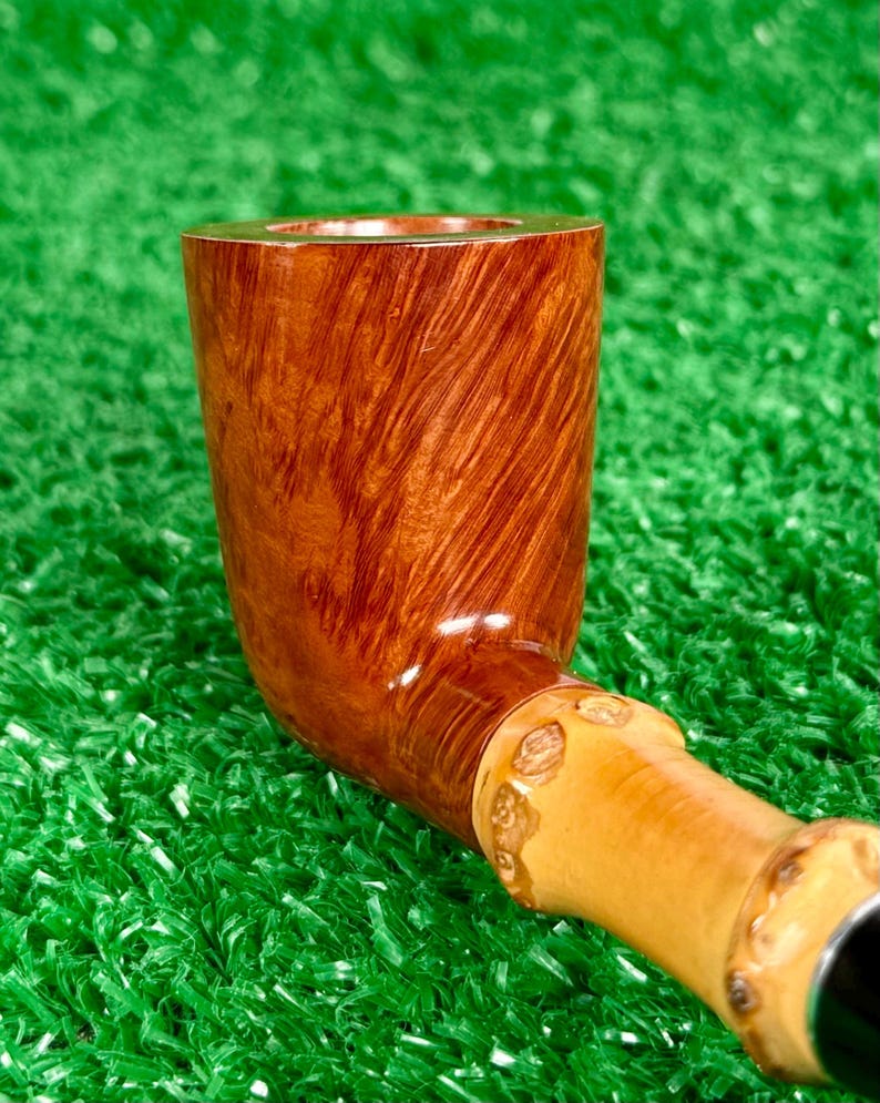 May include: A handcrafted tobacco pipe with a curved, polished wooden bowl and a light-colored stem. The bowl has a rich, reddish-brown hue with visible wood grain. The stem features decorative carvings and a black mouthpiece.