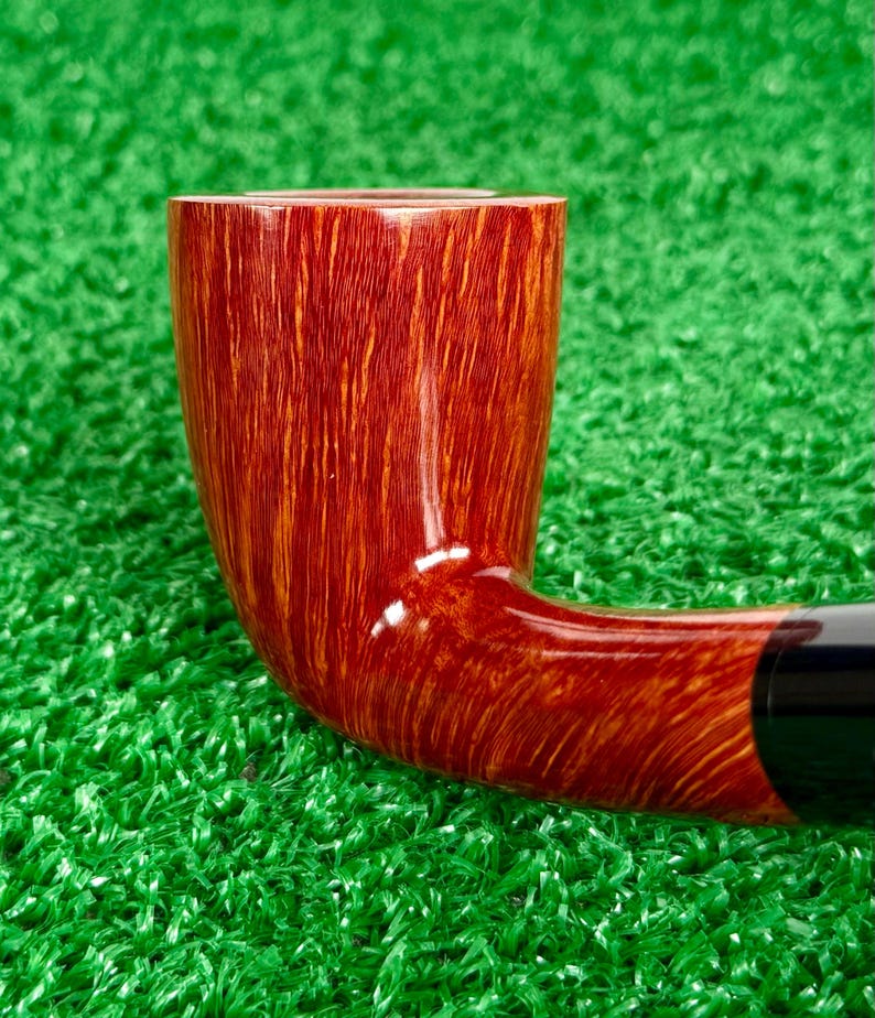 May include: A tobacco pipe featuring a polished, reddish-brown wooden bowl with a prominent vertical grain pattern. The bowl transitions into a smooth, curved shank that connects to a dark, solid-colored stem, showcasing a glossy finish.