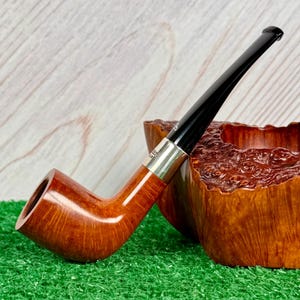 Puede incluir: Una pipa de tabaco con un hornillo de madera marr&oacute;n claro pulido y una boquilla negra larga y delgada. Una banda de metal de color plateado conecta el hornillo con la boquilla. La pipa est&aacute; junto a un objeto de madera texturizado de color marr&oacute;n rojizo oscuro.