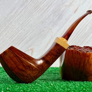 Pode incluir: Um cachimbo de tabaco de madeira castanha polida com um fornilho curvo e liso e uma piteira longa e fina. O cachimbo inclui um anel decorativo de madeira clara na jun&ccedil;&atilde;o entre a piteira e o fornilho, exibindo um veio de madeira natural.
