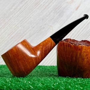 May include: A smooth, light brown wooden tobacco pipe with a long, tapered black stem. The bowl features the engraved text CASTELLO CASTELLO. It is positioned next to a large, textured, reddish-brown wooden block with a rough, natural surface finish.