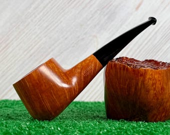 May include: A smooth, light brown wooden tobacco pipe with a long, tapered black stem. The bowl features the engraved text CASTELLO CASTELLO. It is positioned next to a large, textured, reddish-brown wooden block with a rough, natural surface finish.