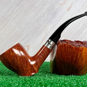 May include: A polished brown wooden tobacco pipe with a smooth, curved grain pattern, featuring a black acrylic stem and a silver-colored metal band connecting the bowl to the stem. The pipe is positioned next to a matching wooden block.