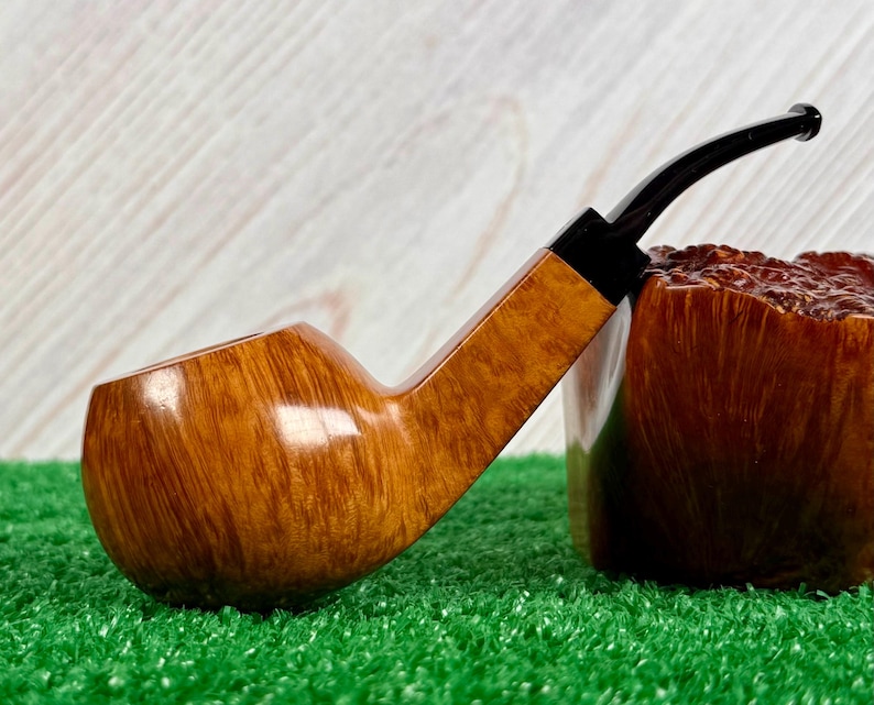 May include: A tobacco pipe featuring a smooth, light brown wooden bowl with a visible grain pattern and a long, slender black stem. The pipe is positioned next to a dark brown, textured wooden block that displays a rough, natural surface finish.