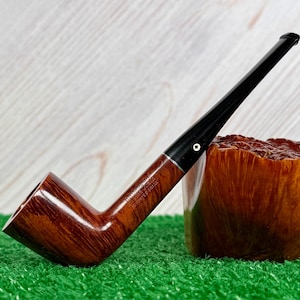 May include: A brown wooden tobacco pipe with a long black stem and a silver-colored metal band. The pipe features a smooth, polished finish and includes the stamped text "BRUYERE GARANTIE" and "IMPORTE D'BRIAR" on the side of the bowl.