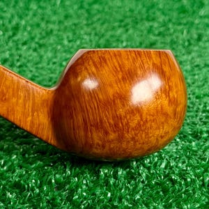 May include: A tobacco pipe bowl crafted from polished light brown wood with a visible natural grain pattern. The bowl features a smooth, rounded shape with a glossy finish that reflects light, showing the intricate texture of the wood grain.