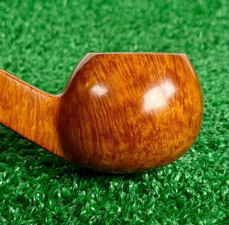 May include: A tobacco pipe bowl crafted from polished light brown wood with a visible natural grain pattern. The bowl features a smooth, rounded shape with a glossy finish that reflects light, showing the intricate texture of the wood grain.