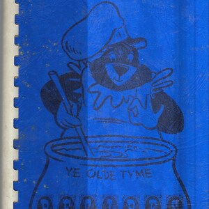 Peut inclure: Une couverture bleue pour un livre de recettes avec un personnage de dessin animé portant un chapeau de chef et tenant une cuillère en bois au-dessus d'une casserole. Le texte sur la couverture est "Ye Olde Tyme Recipes" et "Compiled by Perry Senior Citizens Council 1973".