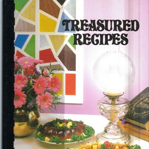 May include: A cookbook titled "Treasured Recipes from Around the World" with a colorful stained glass window in the background. The book is open to a page showing a recipe for a salad with olives, tomatoes, and parsley.