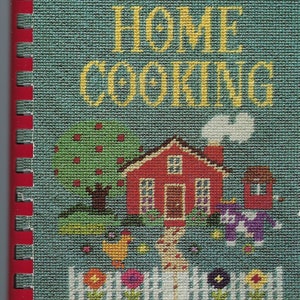 May include: A red and white cookbook with a blue cover and a stitched design of a house, a tree, and flowers. The title is "Our Best Home Cooking" and the text at the bottom reads "Kickapoo Sidekicks Danville, Illinois".