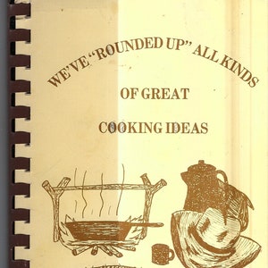Puede incluir: Una portada de libro de cocina vintage con fondo marrón y una ilustración marrón de una fogata con una olla en la parte superior, una cafetera y una taza. El texto de la portada dice "We've "Rounded Up" All Kinds Of Great Cooking Ideas" y "The Phoenix Get-Together Extension Club".