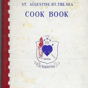 Può includere: La copertina di un libro di cucina vintage intitolato "St. Augustine-By-The-Sea Cook Book" con uno stemma blu e bianco che presenta un cuore, frecce e le parole "Veritas" e "St. Augustine". Il testo "Compilato dalle donne della chiesa" è stampato sotto lo stemma.