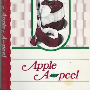May include: A red apple with a green leaf on top is being peeled with a silver knife. The apple is surrounded by a green oval frame. The text "Apple A-peel" is written in red cursive letters below the frame.